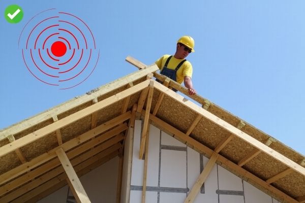 A construction worker reinforcing a roof for seismic resistance. The worker is installing steel reinforcements and securing wooden beams on the roof structure of a residential building. The focus